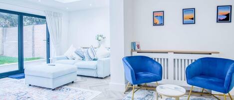Una sala de estar moderna con un sofá blanco, sillones azules y una mesa de centro. Hay tres cuadros enmarcados en la pared y un ventanal que proporciona luz natural.