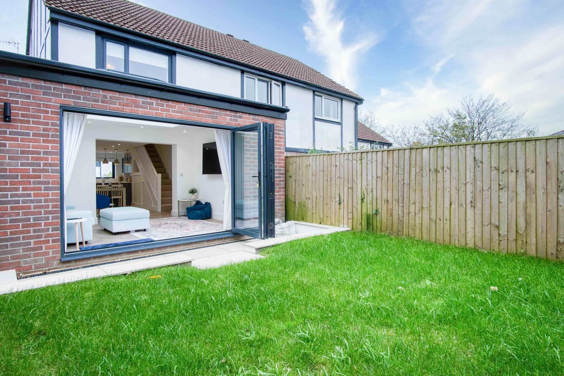 A modern house with a brick facade, a large glass door leading to a living area with a sofa and a coffee table, and a wooden fence in the backyard.
