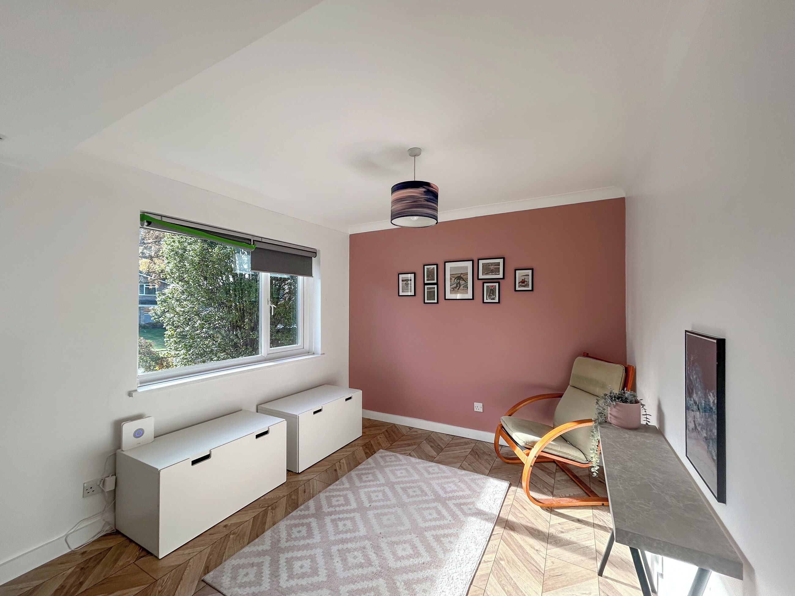 A room with a window, a ceiling fan, a wall with framed pictures, a chair, a desk, and a patterned rug.