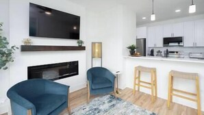 A modern living room with a fireplace, a TV mounted on the wall, and a kitchen area with white cabinets and a black refrigerator. - Brick City Lofts Downtown Ocala Unit 303 (Ocala)