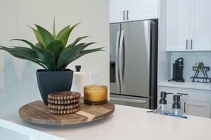 A modern kitchen with a stainless steel refrigerator, a potted plant, and a wooden serving tray with coasters and a candle. - Brick City Lofts Downtown Ocala Unit 303 (Ocala)