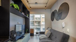 A modern living room with a sofa, a TV, and a fan. There are shelves with plants and a window with a view of buildings. - Laguna 430 (São Paulo)