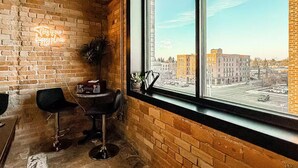 Quarto de canto com parede de tijolos, um letreiro de neon, uma planta em vaso, um rádio antigo e vista para a cidade através de uma grande janela.