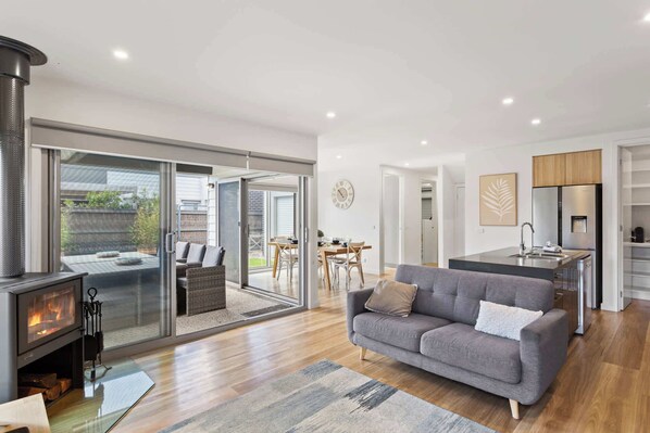 A modern living room with a fireplace, a sofa, a dining area, and a kitchen with a refrigerator. - Golf Course Front Stylish Townhouse w/ Pool Access (Cowes)