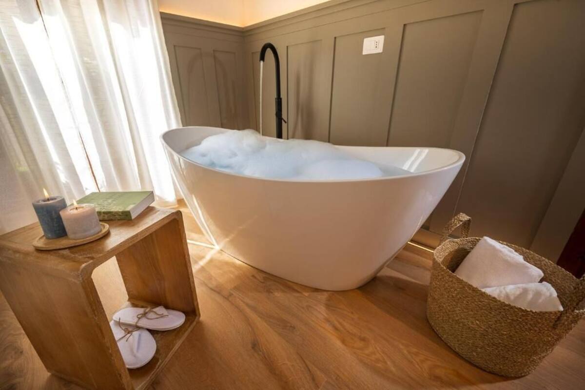 A modern bathroom with a freestanding bathtub filled with bubbles, a wooden side table with candles and slippers, and a woven basket with towels.