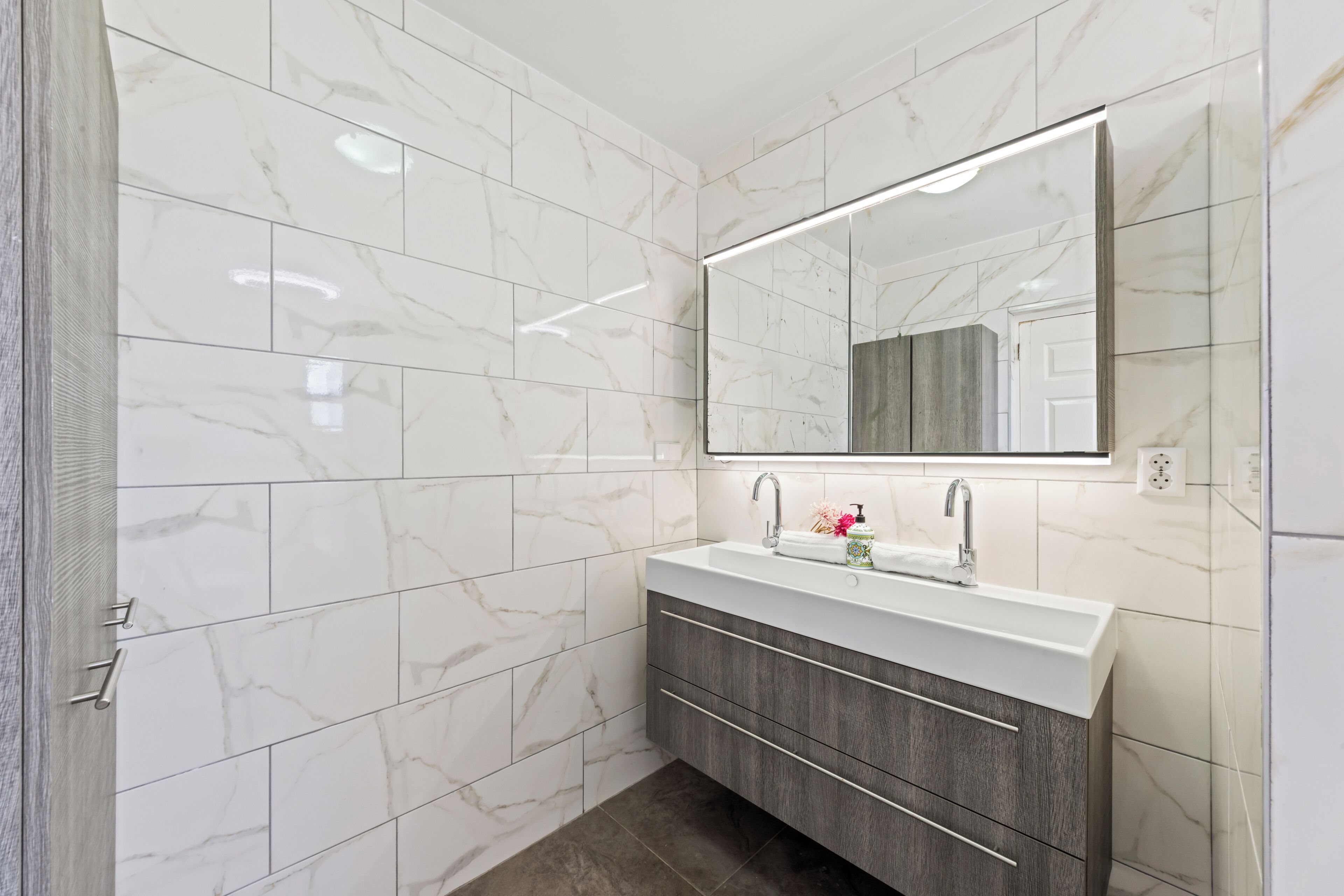 Une salle de bain avec des carreaux de marbre blanc, un lavabo blanc doté d’un robinet moderne, un grand miroir et un meuble-lavabo en bois.