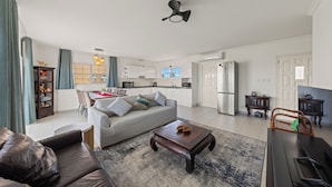 Una sala de estar con sofá, mesa de centro y televisor. Se observa una cocina al fondo con armarios y un refrigerador.