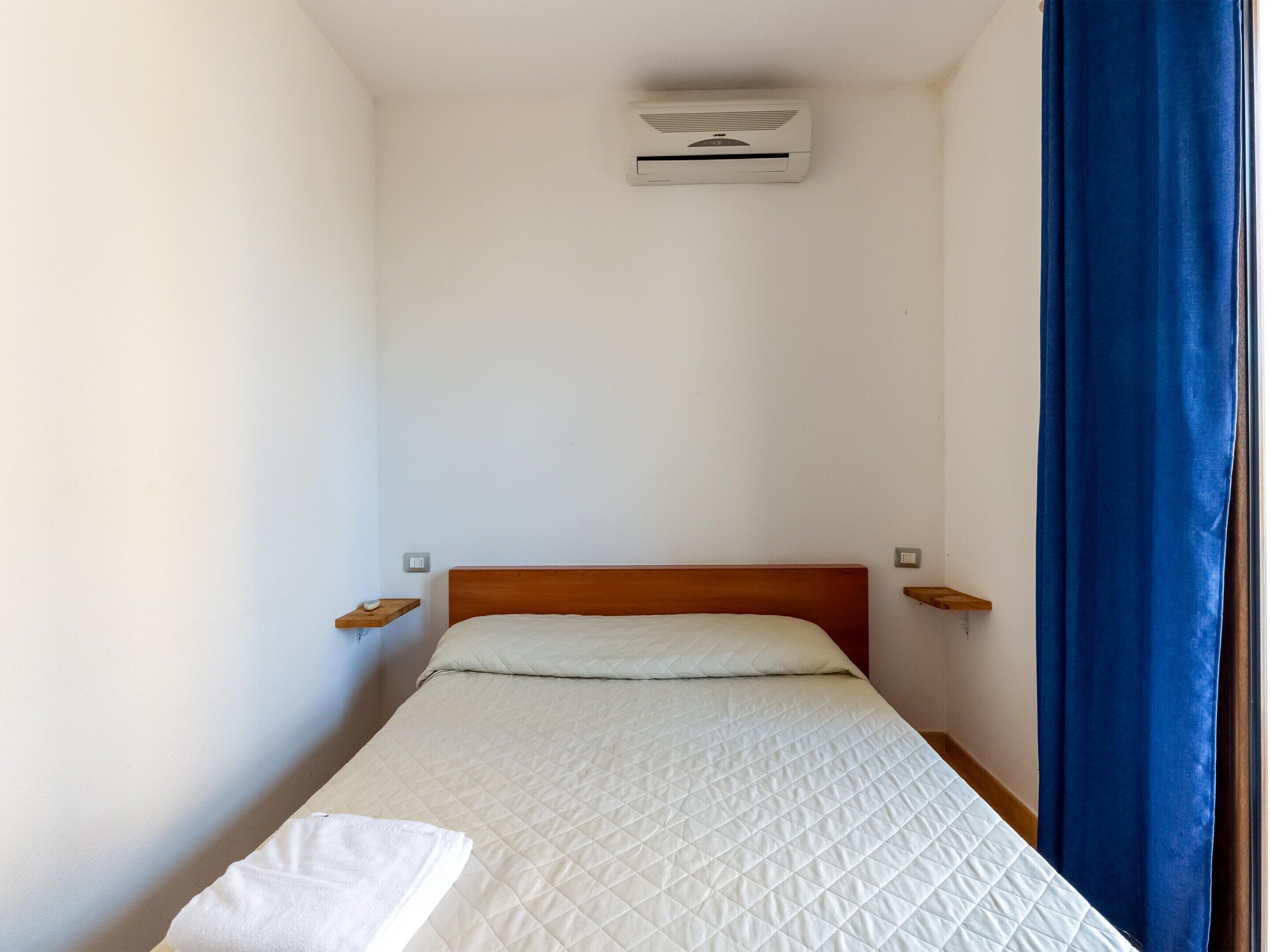 Una cama individual con cabecera de madera, colchón blanco acolchado y una mesita de noche con lámpara.