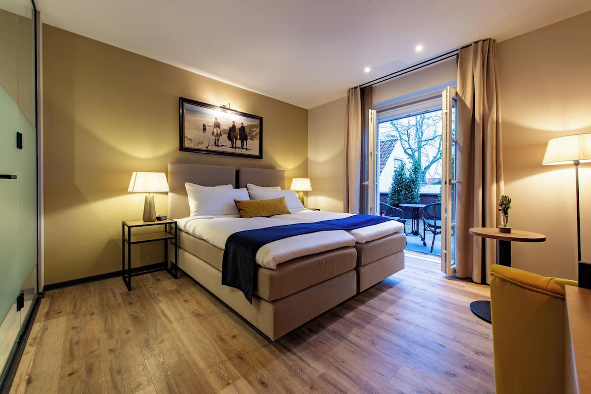 Une chambre d’hôtel moderne avec un grand lit, deux tables de chevet avec des lampes, un tableau au-dessus du lit, une porte-fenêtre coulissante donnant sur un balcon et une petite table avec un vase.