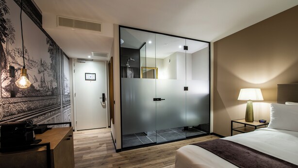 Une chambre d’hôtel moderne avec un bureau en bois, un lit recouvert d’un plaid marron, une table de chevet avec une lampe et un grand miroir.