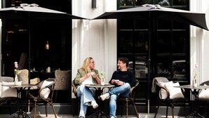 Un couple est assis à une table avec des chaises, dans un café en plein air, protégé du soleil par un grand parasol.