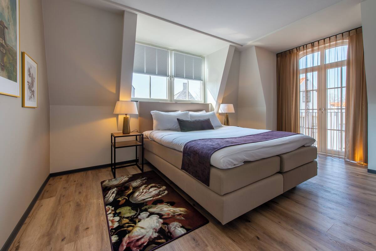 Une chambre moderne avec un grand lit, une fenêtre avec des stores, une table de chevet et une lampe sur pied.