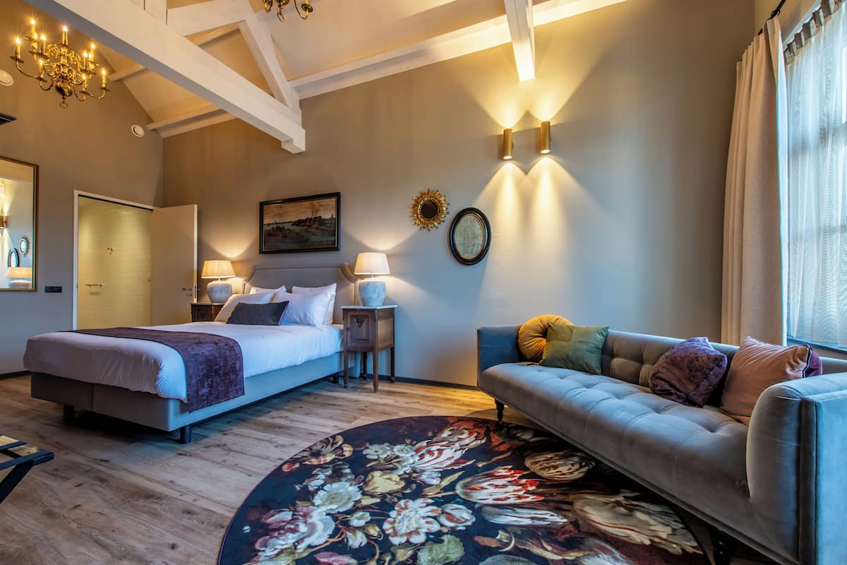 Une chambre d’hôtel avec un lit, un canapé, un tapis rond, un lustre, des appliques murales et un tableau.