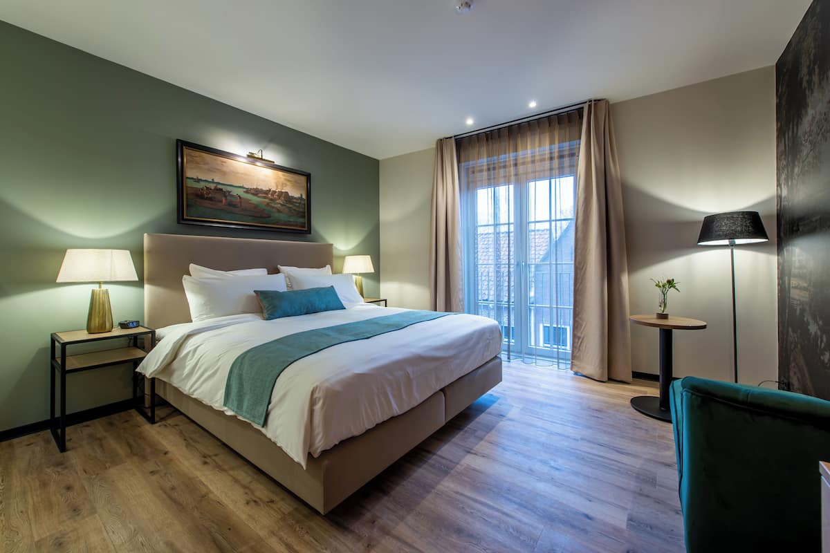 Une chambre d’hôtel avec un grand lit, deux tables de chevet avec des lampes, une petite table ronde avec une lampe, un fauteuil vert et une grande fenêtre avec des rideaux.