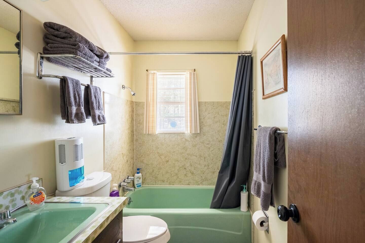 Un baño con una bañera verde, un lavamanos blanco, un inodoro, un perchero con toallas dobladas, una cortina de ducha y una ventana con persianas.