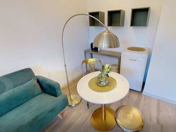 A modern living room with a teal sofa, a round white table with a gold base, a floor lamp, and a white mini-fridge. - 739A goldfinger+suites I Apartment Unique 1 (Lana)