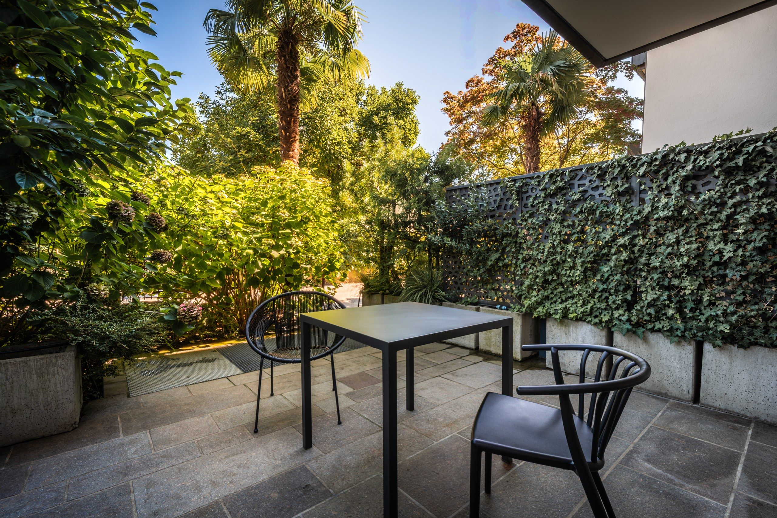 Un patio exterior con mesa y sillas, rodeado de abundante vegetación y un muro decorativo.