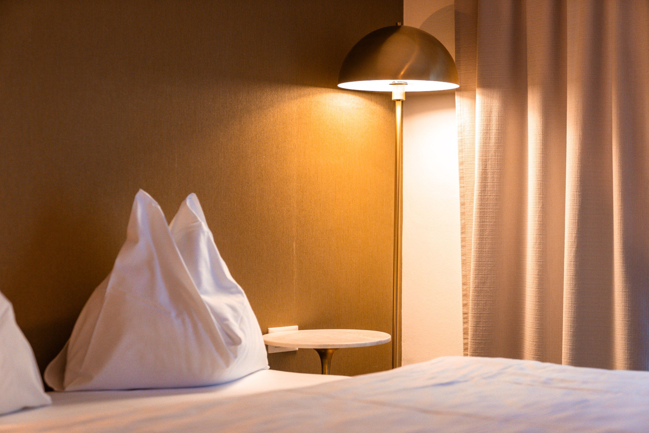 Een hotelkamer met een bed, kussens, een klein rond tafeltje, een vloerlamp en gordijnen.
