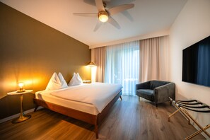 A hotel room with a bed, a chair, a desk, a lamp, a ceiling fan, and a large window with curtains.