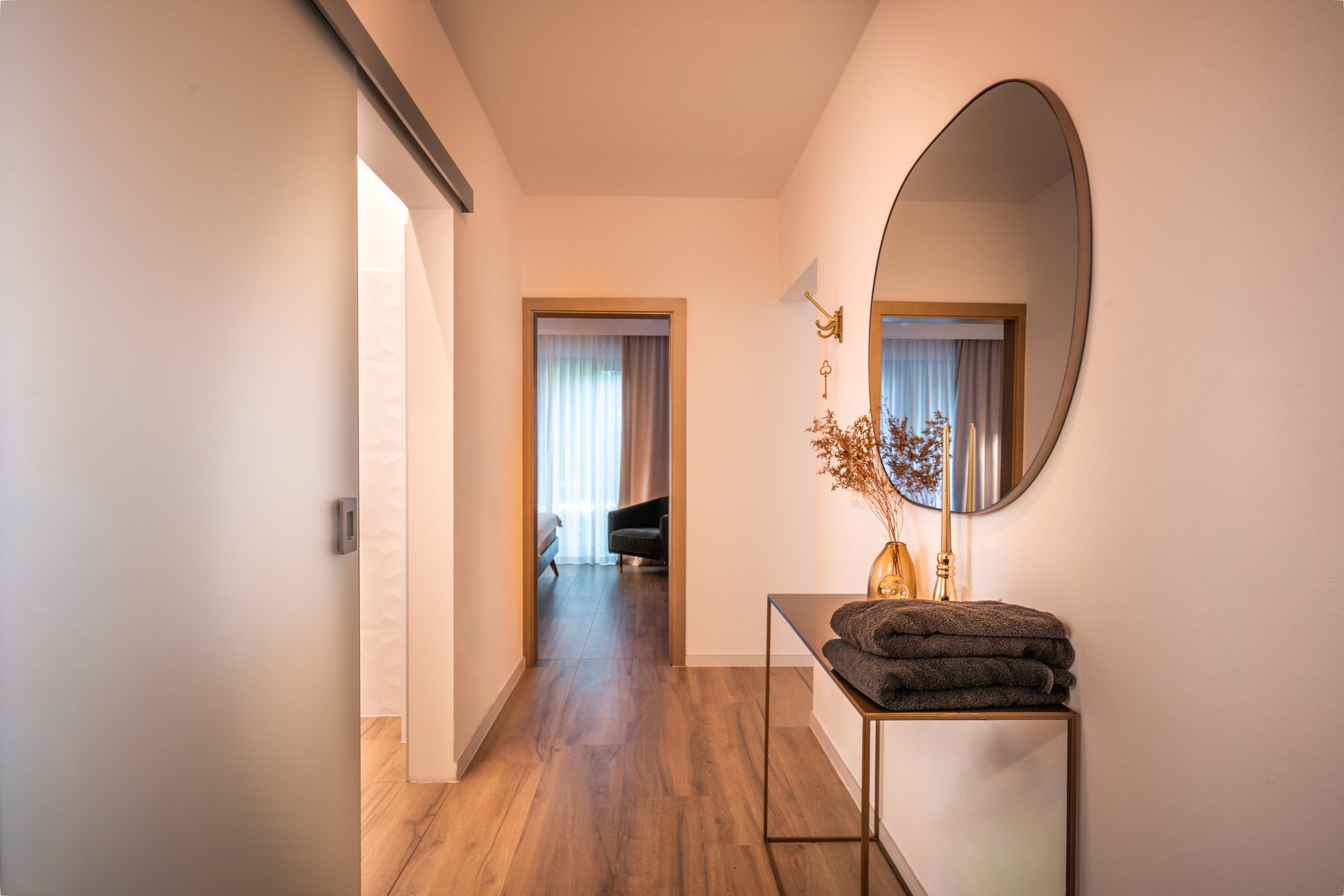 A hallway with wooden flooring, a bed with a headboard, a chair, and a table with a mirror and a vase with dried flowers.