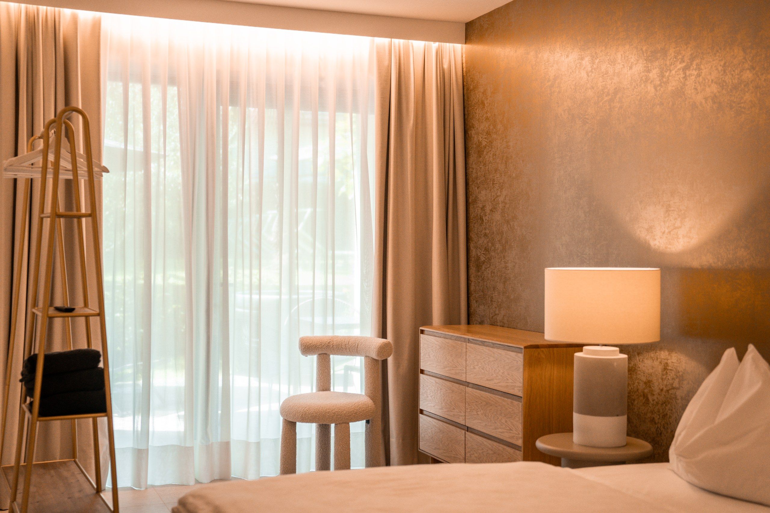 Ein Hotelzimmer mit einem Bett, einem Nachttisch mit Lampe, einem Sessel, einer Kommode und einem Fenster mit Vorhängen.