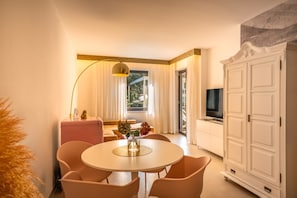 Ein Hotelzimmer mit einem runden Esstisch, Stühlen, einem Fernseher, einem Schrank und einem Fenster mit Vorhängen.