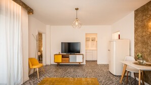 A modern living room with a flat-screen TV, a yellow chair, a white dining table, and a patterned rug. - 739F goldfinger+suites I Apartment Unique 15 (Lana)