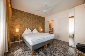 A hotel room with a bed, a ceiling fan, a patterned floor, and a bathroom visible through an open door. - 739F goldfinger+suites I Apartment Unique 15 (Lana)