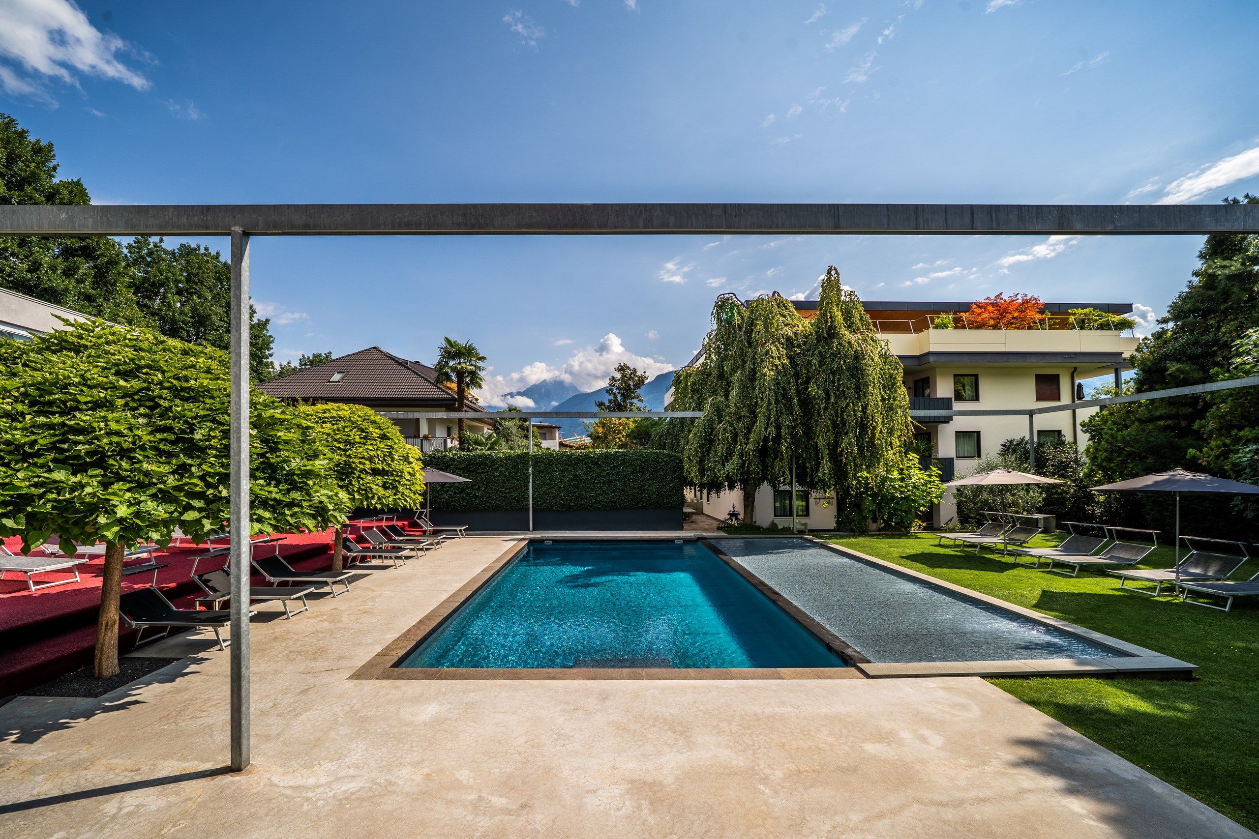 Une piscine rectangulaire à l’eau d’un bleu limpide, entourée d’une terrasse en béton. On y trouve des chaises longues et des parasols, ainsi qu’une haie avec un grand arbre en arrière-plan.