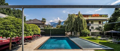 A rectangular swimming pool with a clear blue water surface, surrounded by a concrete deck. There are lounge chairs and umbrellas on the deck, and a hedge with a tall tree in the background.