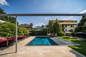 A rectangular swimming pool with a clear blue water surface, surrounded by a concrete deck. There are lounge chairs and umbrellas on the deck, and a hedge with a tall tree in the background. - 739F goldfinger+suites I Apartment Unique 15 (Lana)