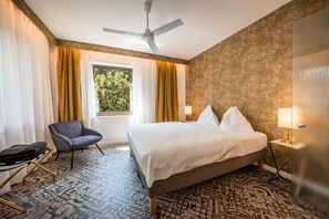 Une chambre d’hôtel avec un lit, une chaise, une fenêtre avec des rideaux, un ventilateur de plafond et un revêtement de sol à motifs.