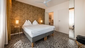 A hotel room with a bed, a ceiling fan, a large mirror, and patterned flooring. - 739F goldfinger+suites I Apartment Unique 15 (Lana)