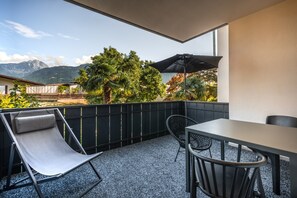 Uma varanda com mesa e cadeiras, um guarda-sol preto e vista para montanhas e árvores.
