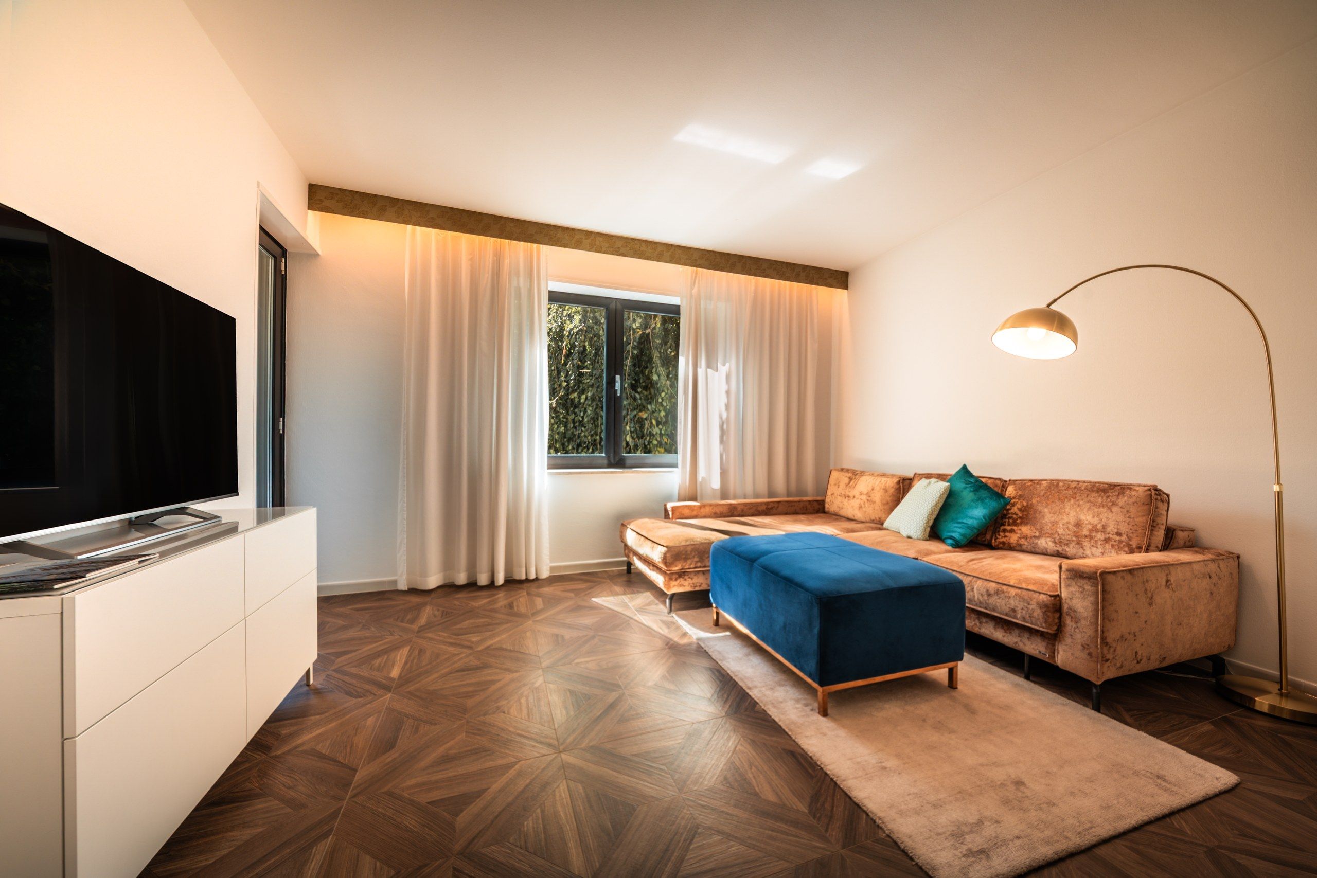Ein modernes Wohnzimmer mit einem Flachbildfernseher, einer Couch, einem blauen Hocker, einer Stehlampe und einem Fenster mit Vorhängen.