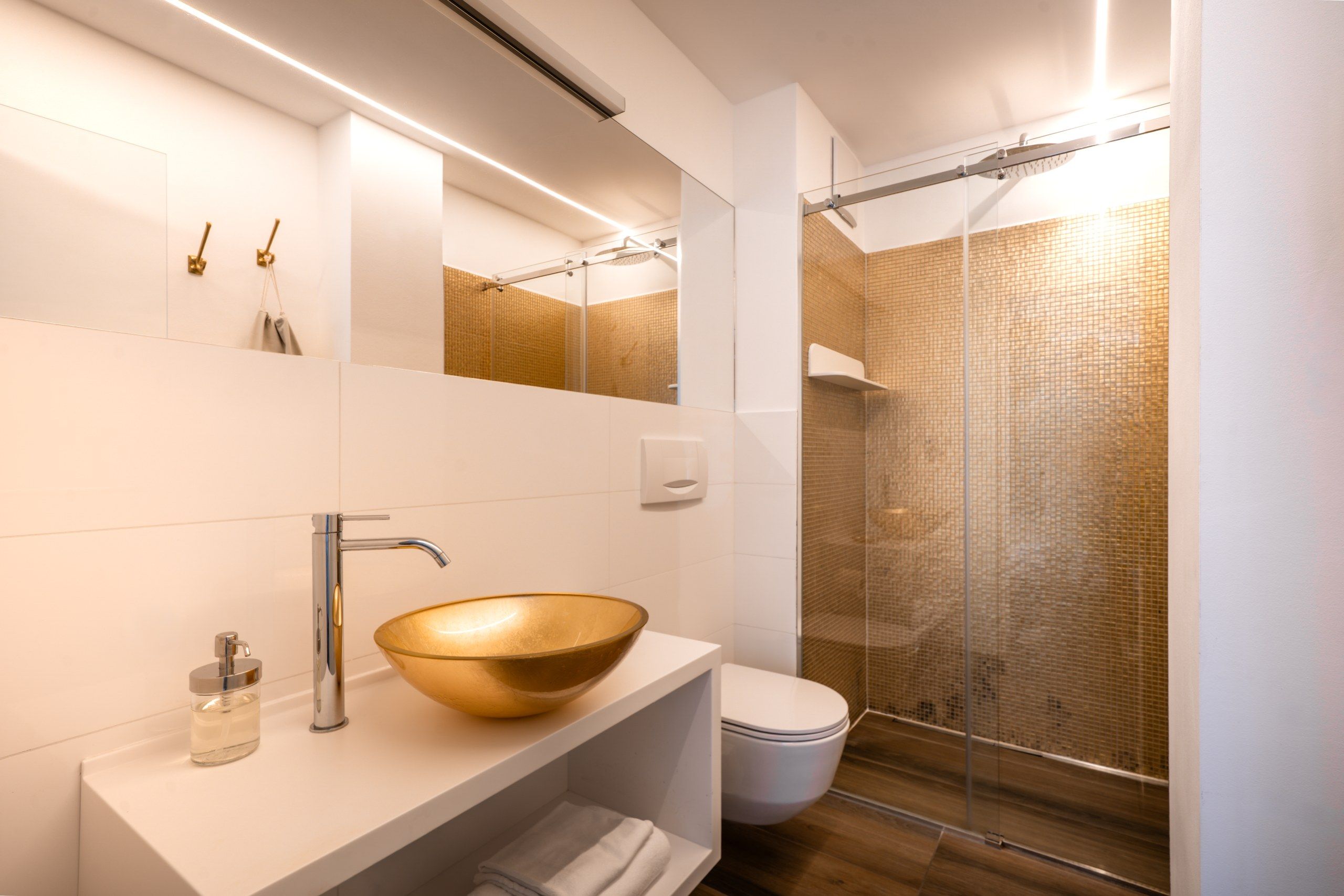 Un bany modern amb un gran mirall, un lavabo daurat, un vàter i una dutxa amb envàs de vidre.