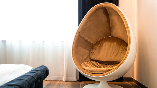 Een moderne kamer met een opvallende, eivormige fauteuil, een bed met een witte dekbedovertrek en een raam met dunne gordijnen.