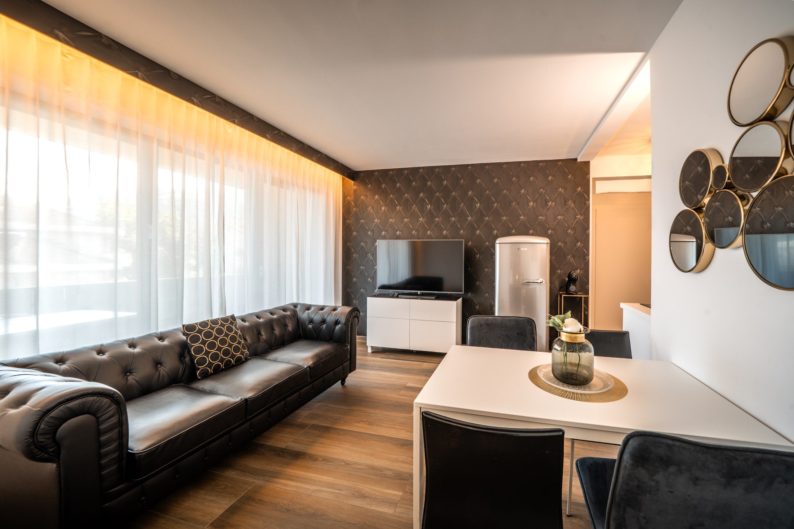 A modern living room with a leather sofa, a flat-screen TV, a coffee machine, and a dining area with a white table and chairs.