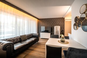 A modern living room with a leather sofa, a flat-screen TV, a coffee machine, and a dining area with a white table and chairs.
