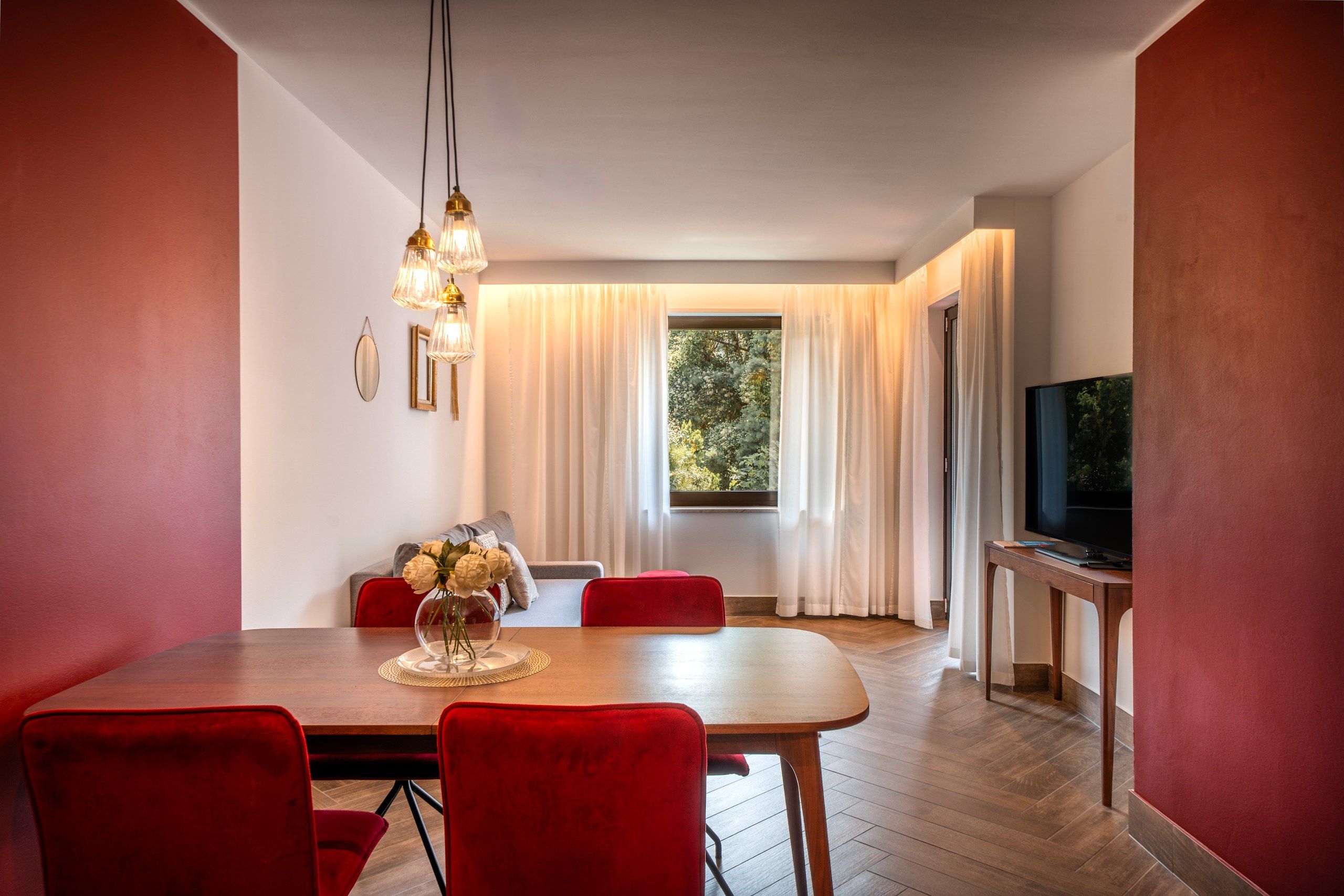 A modern dining area with a wooden table, red chairs, a vase of flowers, and a television on a wooden stand.