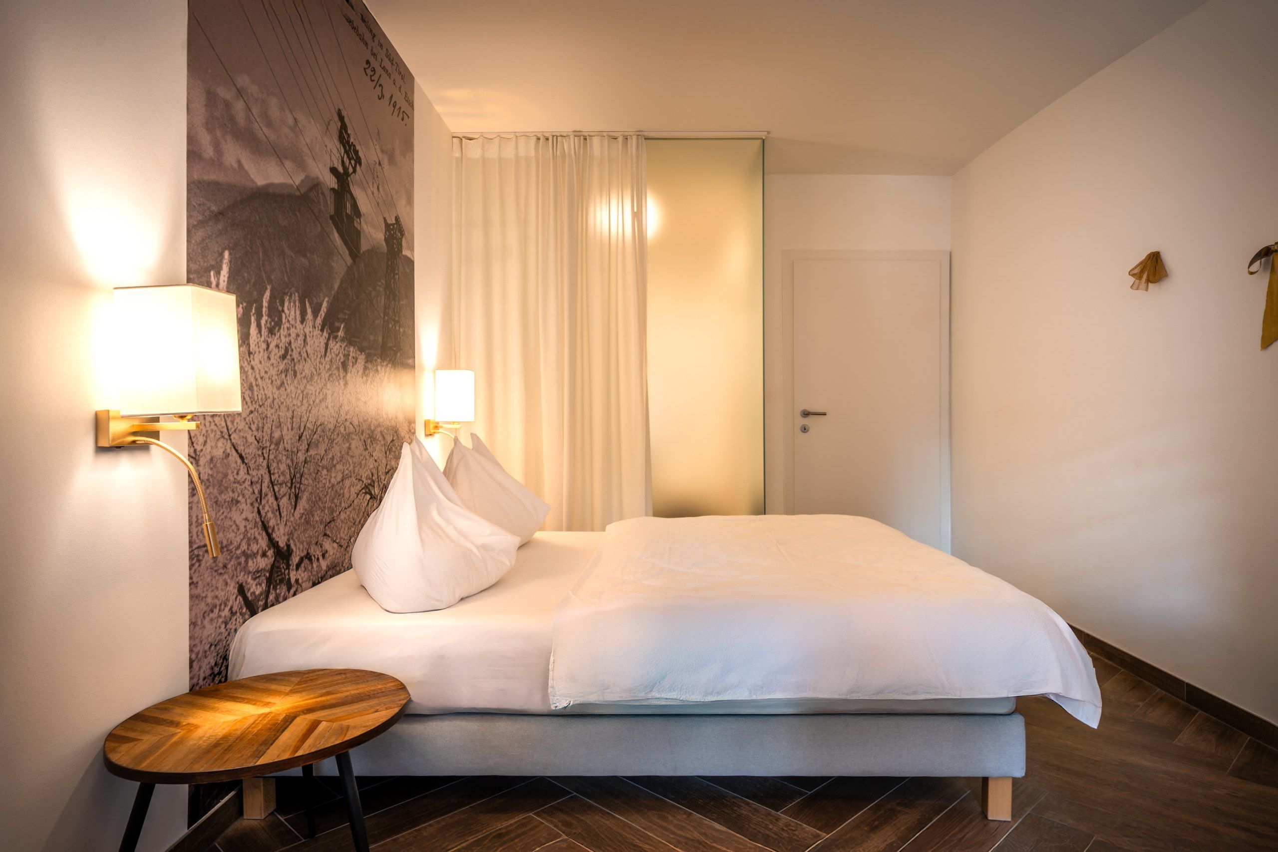 A hotel room with a bed, a small wooden table, two wall-mounted lamps, and a door.