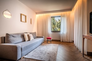Ein modernes Wohnzimmer mit einem grauen Sofa, einem kleinen roten Hocker, einem Holztisch und einem großen Fenster mit durchsichtigen Vorhängen.