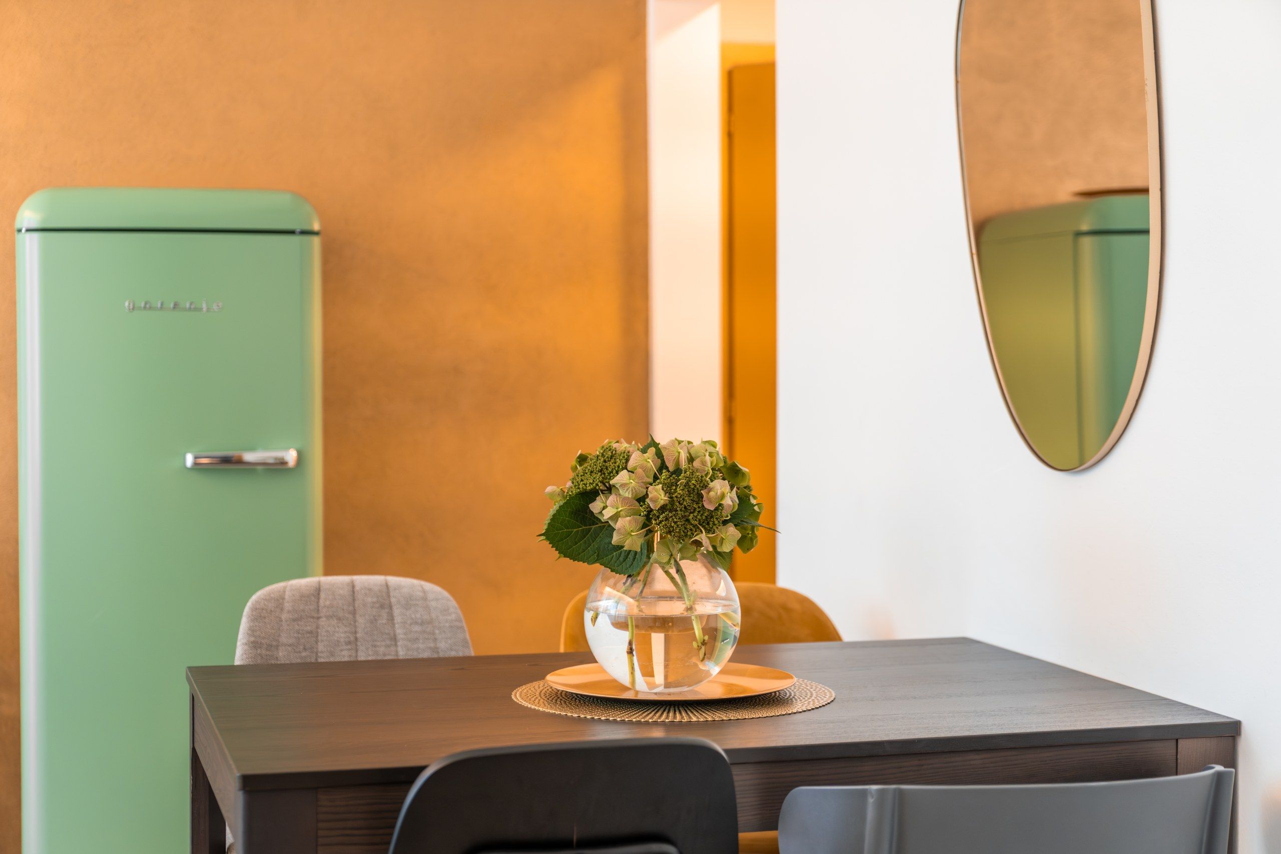 Un coin repas comprenant une table, des chaises, un vase avec des fleurs, un réfrigérateur vert et un miroir au mur.