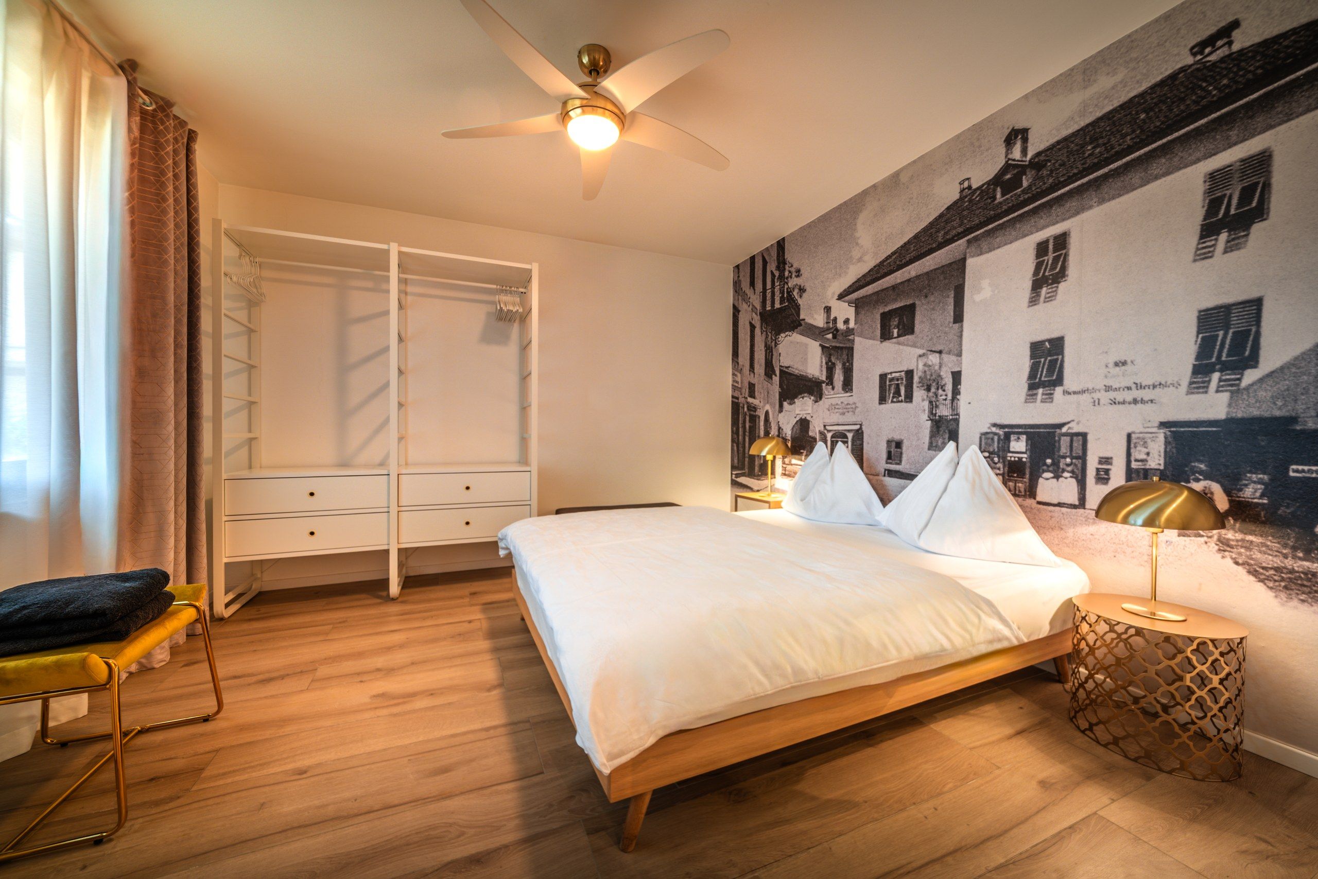 Quarto com cama, guarda-roupa, ventoinha no teto, mesa de cabeceira e parede com mural de paisagem urbana em preto e branco.