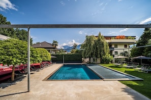 A rectangular swimming pool with a clear blue water surface, surrounded by a concrete deck. There are lounge chairs and umbrellas on the deck, and a hedge with a tall tree in the background.