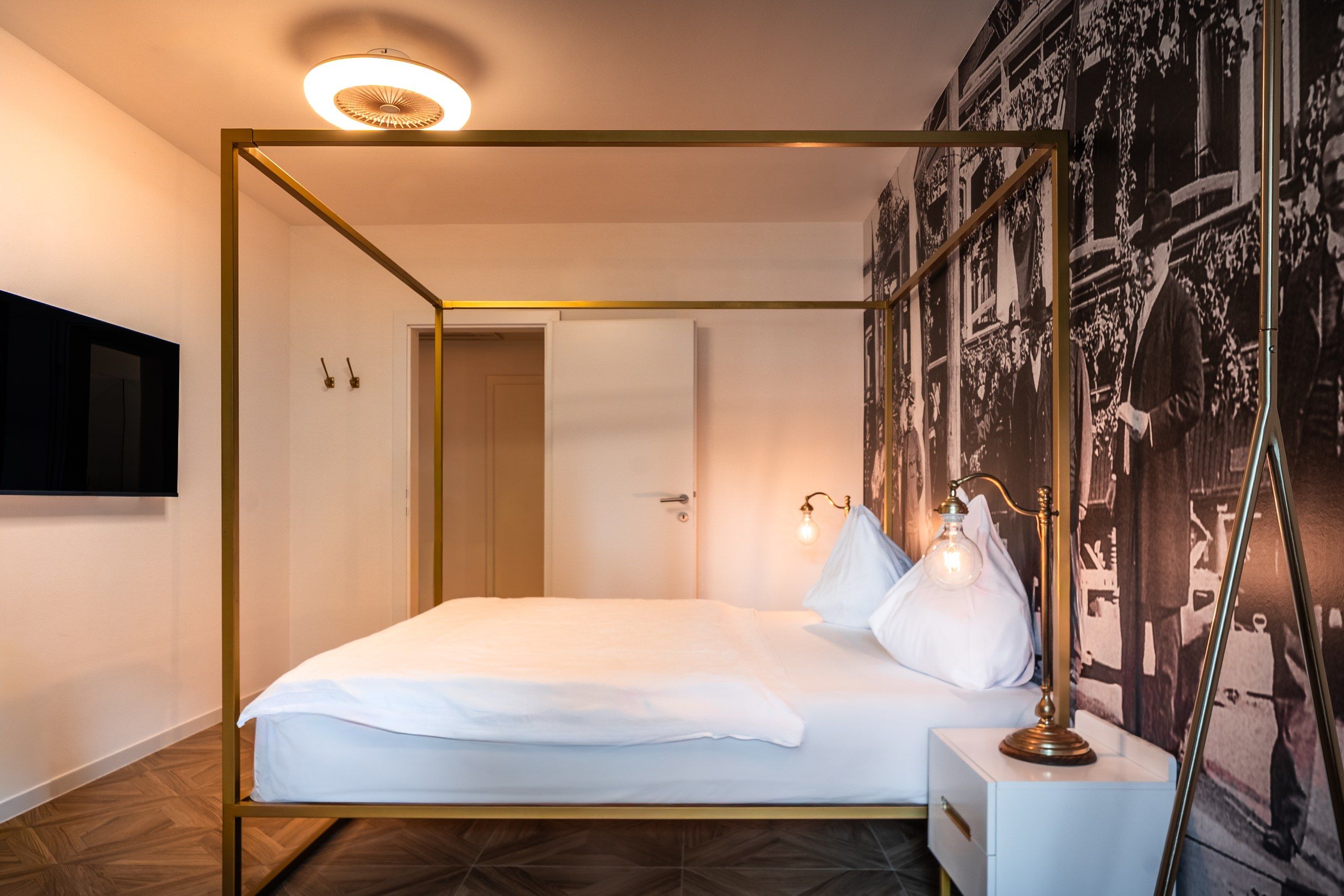 Un lit à baldaquin avec un matelas et des oreillers blancs, une table de chevet avec une lampe et un téléviseur fixé au mur.