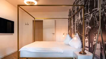 Een hemelbed met een witte matras en kussens, een nachtkastje met een lamp en een aan de muur bevestigde televisie.