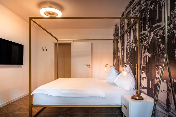 Una cama con dosel, un colchón y almohadas blancos, una mesita de noche con una lámpara y un televisor montado en la pared.