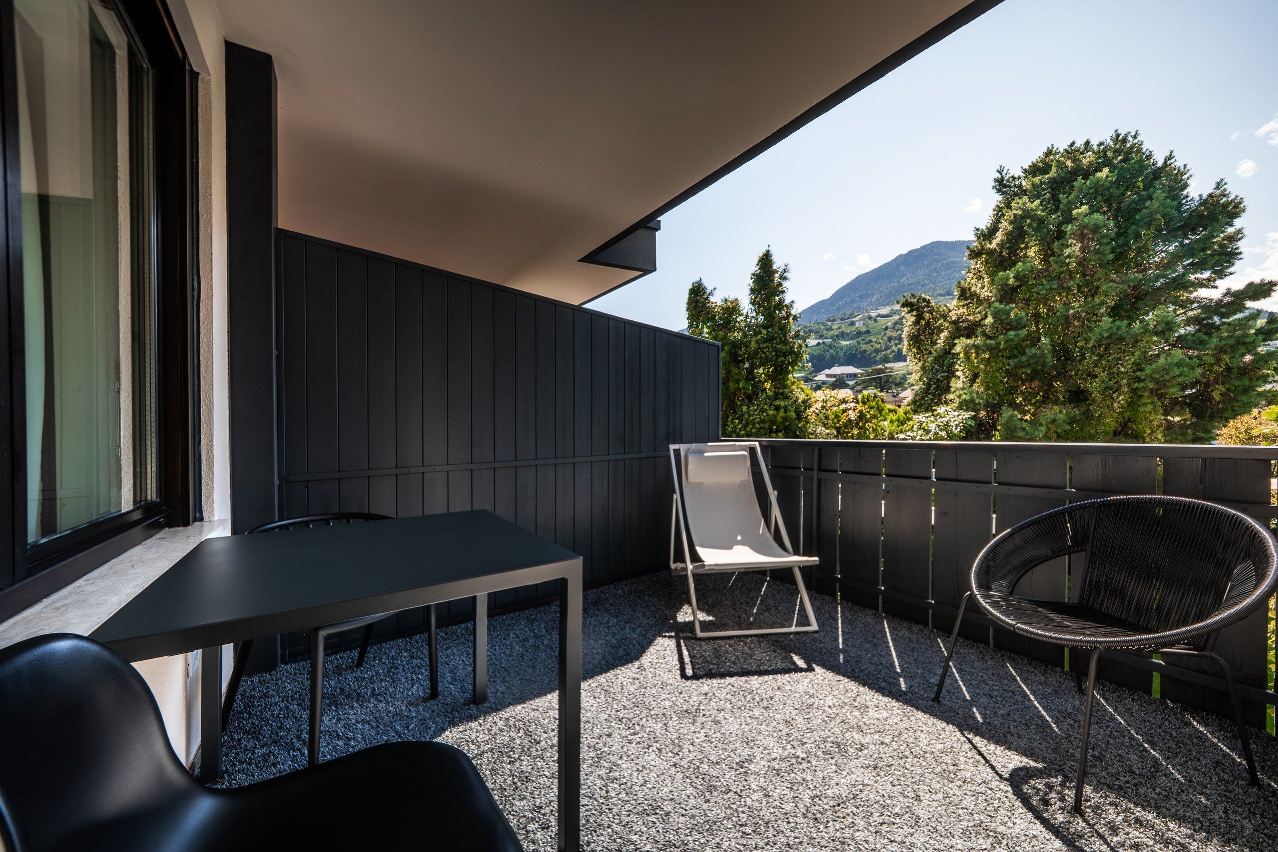 Een balkon met een tafel en stoelen, uitzicht op bomen en bergen, en een houten hek.