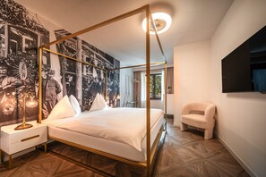 A four-poster bed with a gold frame, a nightstand with a lamp, and a wall with a large black-and-white photograph. - 739J goldfinger+suites I Apartment Individual 22 (Lana)